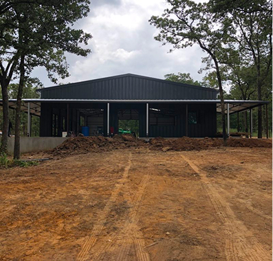 Black metal building under construction with dirt yard and trees.